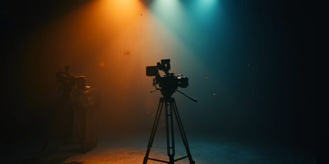 Empty television studio with dimmed lights and dormant camera equipment on tripod, bathed in warm amber and deep blue tones