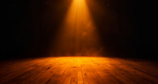 Empty theater stage illuminated by a single amber spotlight amid darkness, wooden floorboards catching the glow at twilight