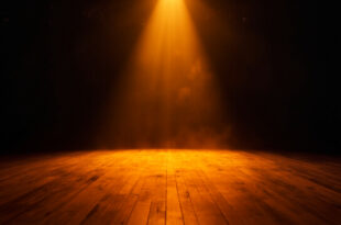 Empty theater stage illuminated by a single amber spotlight amid darkness, wooden floorboards catching the glow at twilight