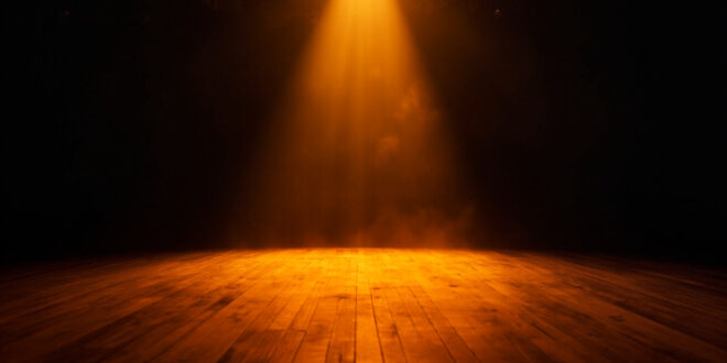 Empty theater stage illuminated by a single amber spotlight amid darkness, wooden floorboards catching the glow at twilight