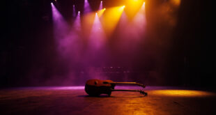 Abandoned acoustic guitar sits alone on dimly lit concert stage, bathed in golden spotlight against deep purple backdrop