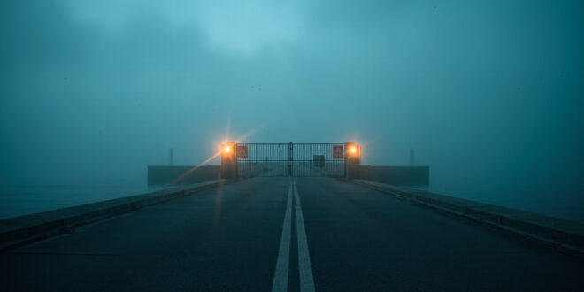 Abandoned coastal border checkpoint at dawn with closed security gate and warning signs overlooking misty waters