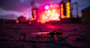 Abandoned syringe on concrete amid empty festival grounds at dusk, with scattered debris near a deserted stage