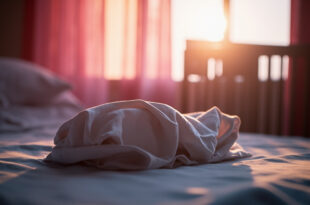 Empty hospital bassinet beside window at dawn, with unopened pink blanket on nearby bed in quiet maternity room