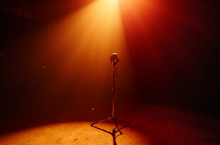A spotlit vintage microphone stands alone in an empty recording studio, bathed in burgundy and gold light, symbolizing loss.