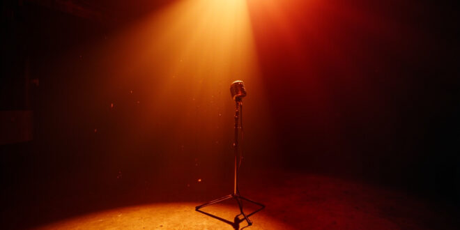 A spotlit vintage microphone stands alone in an empty recording studio, bathed in burgundy and gold light, symbolizing loss.
