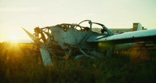 Damaged glider aircraft wreckage on empty airfield grass with hospital building visible in background