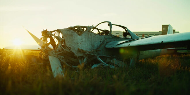 Damaged glider aircraft wreckage on empty airfield grass with hospital building visible in background
