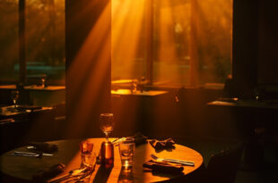 Empty round table with multiple place settings and one highlighted chair in a dimly lit restaurant, evoking solitude.
