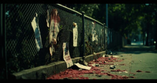 Red paint stains on concrete ground near chain-link fence with torn paper fragments in urban park setting