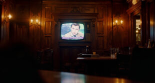Television screen mounted on wooden pub wall displaying quiz show set with warm amber lighting throughout interior