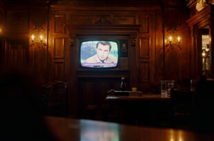 Television screen mounted on wooden pub wall displaying quiz show set with warm amber lighting throughout interior