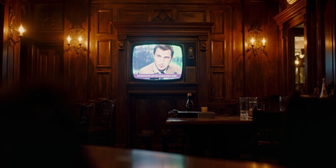 Television screen mounted on wooden pub wall displaying quiz show set with warm amber lighting throughout interior