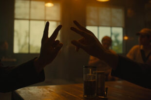 Hands forming sign language gesture above restaurant table with warm lighting and wooden dining tables in soft focus.