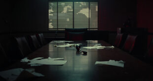 Empty conference room with microphone on wooden table, papers scattered, venetian blind shadows across chairs.