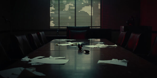 Empty conference room with microphone on wooden table, papers scattered, venetian blind shadows across chairs.