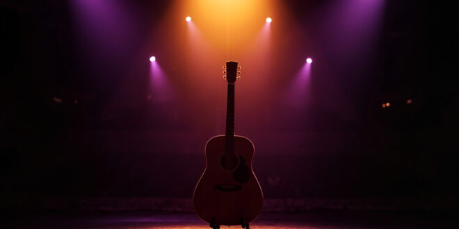 Acoustic guitar on stand under amber spotlight on empty concert stage with purple shadows