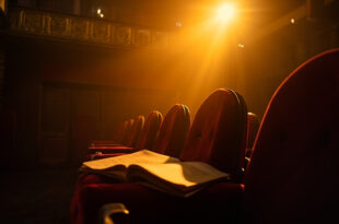 Abandoned Broadway theater with empty seats facing a vacant stage, vintage script resting on a seat in dim burgundy and amber light.