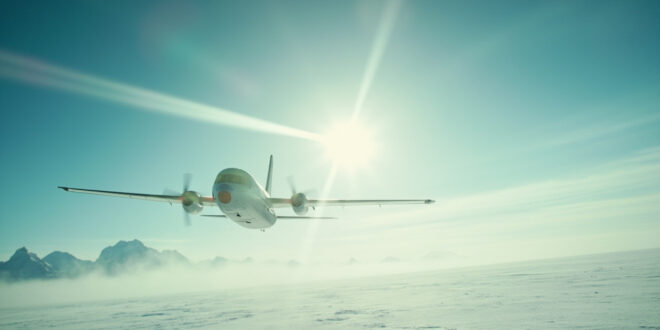 Solitary aircraft flying over snowy Alaskan wilderness approaching distant airport under vast arctic blue sky