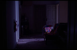 Dark hallway with closed door, colorful fabric draped nearby, scattered personal belongings on floor.