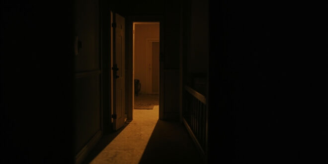 Dark hallway with bedroom door slightly open, warm light filtering through the gap into shadowy corridor.