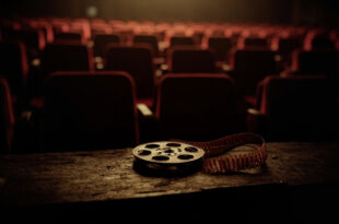 Vintage film reel sits on wooden surface in dimly lit abandoned movie theater with rows of empty red seats