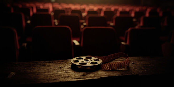 Vintage film reel sits on wooden surface in dimly lit abandoned movie theater with rows of empty red seats