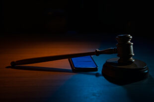 Wooden gavel beside glowing smartphone screen in dark courtroom setting with dramatic lighting