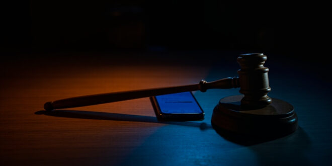 Wooden gavel beside glowing smartphone screen in dark courtroom setting with dramatic lighting