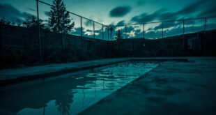 Empty outdoor swimming pool surrounded by metal fencing and warning barriers at twilight