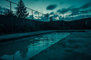 Empty outdoor swimming pool surrounded by metal fencing and warning barriers at twilight