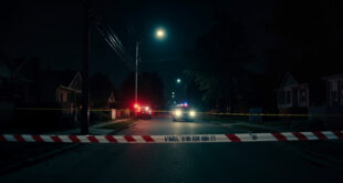 Red and white police tape stretched across empty residential street with emergency vehicles in background