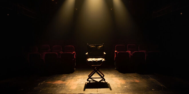 Empty Broadway theater stage with single director's chair under spotlight, red velvet seats in shadows