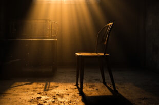 Empty metal chair beside open birdcage in dimly lit underground chamber, illuminated by warm amber light
