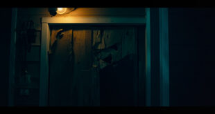 Bullet-damaged wooden front door of suburban home with security camera and amber porch lighting in darkness