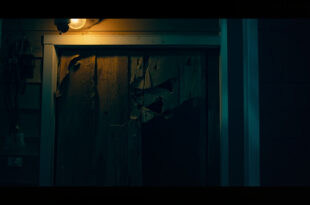 Bullet-damaged wooden front door of suburban home with security camera and amber porch lighting in darkness