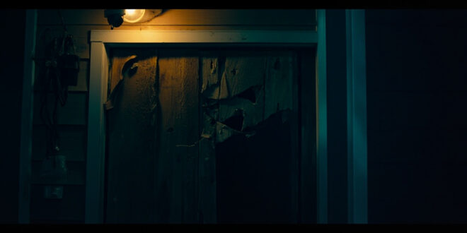 Bullet-damaged wooden front door of suburban home with security camera and amber porch lighting in darkness