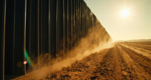 Tall black-painted border wall structure spanning across an empty desert landscape in harsh sunlight