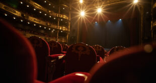 Empty ornate theater with red velvet seats and gold accents facing a darkened stage with closed curtains in a grand concert hall