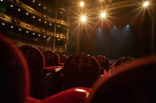 Empty ornate theater with red velvet seats and gold accents facing a darkened stage with closed curtains in a grand concert hall