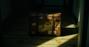 Weathered brown leather suitcase positioned near doorway in dramatic lighting with long shadows on wooden floor