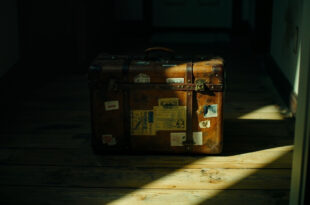 Weathered brown leather suitcase positioned near doorway in dramatic lighting with long shadows on wooden floor