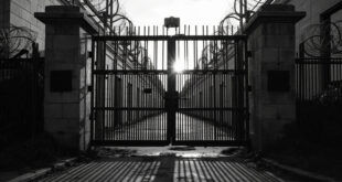 Heavy iron prison gates with concrete walls and razor wire against overcast sky