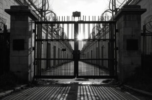 Heavy iron prison gates with concrete walls and razor wire against overcast sky