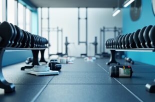 Heavy dumbbells and protein containers arranged on black rubber gym flooring under industrial lighting.