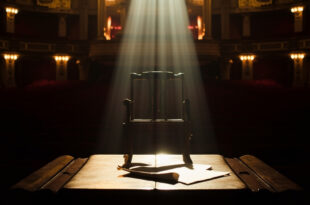 Empty Broadway theater stage with spotlight on vacant chair, script visible on podium, red velvet seats in background
