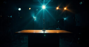 Empty television studio with microphone on desk under dramatic lighting surrounded by cameras and production equipment