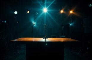 Empty television studio with microphone on desk under dramatic lighting surrounded by cameras and production equipment