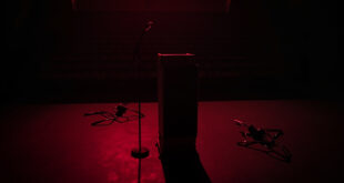 Dark university auditorium with single microphone on podium, empty chairs scattered around, dramatic shadows