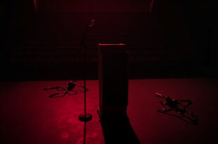 Dark university auditorium with single microphone on podium, empty chairs scattered around, dramatic shadows